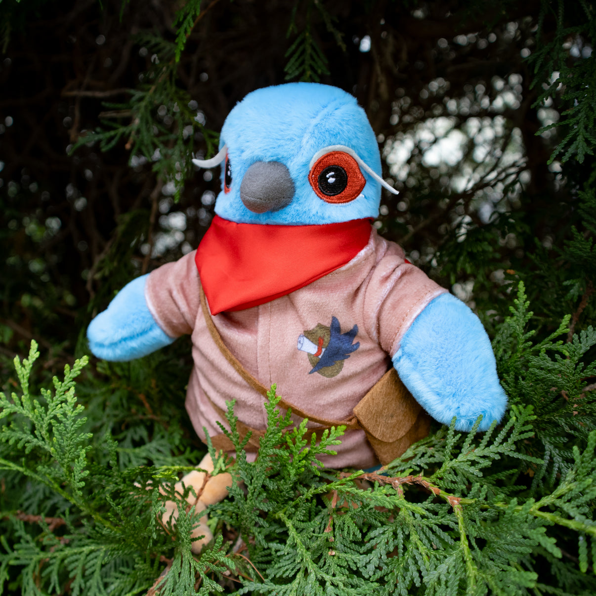 Plush toy resembling a bird with a blue head and red scarf sitting among green foliage.
